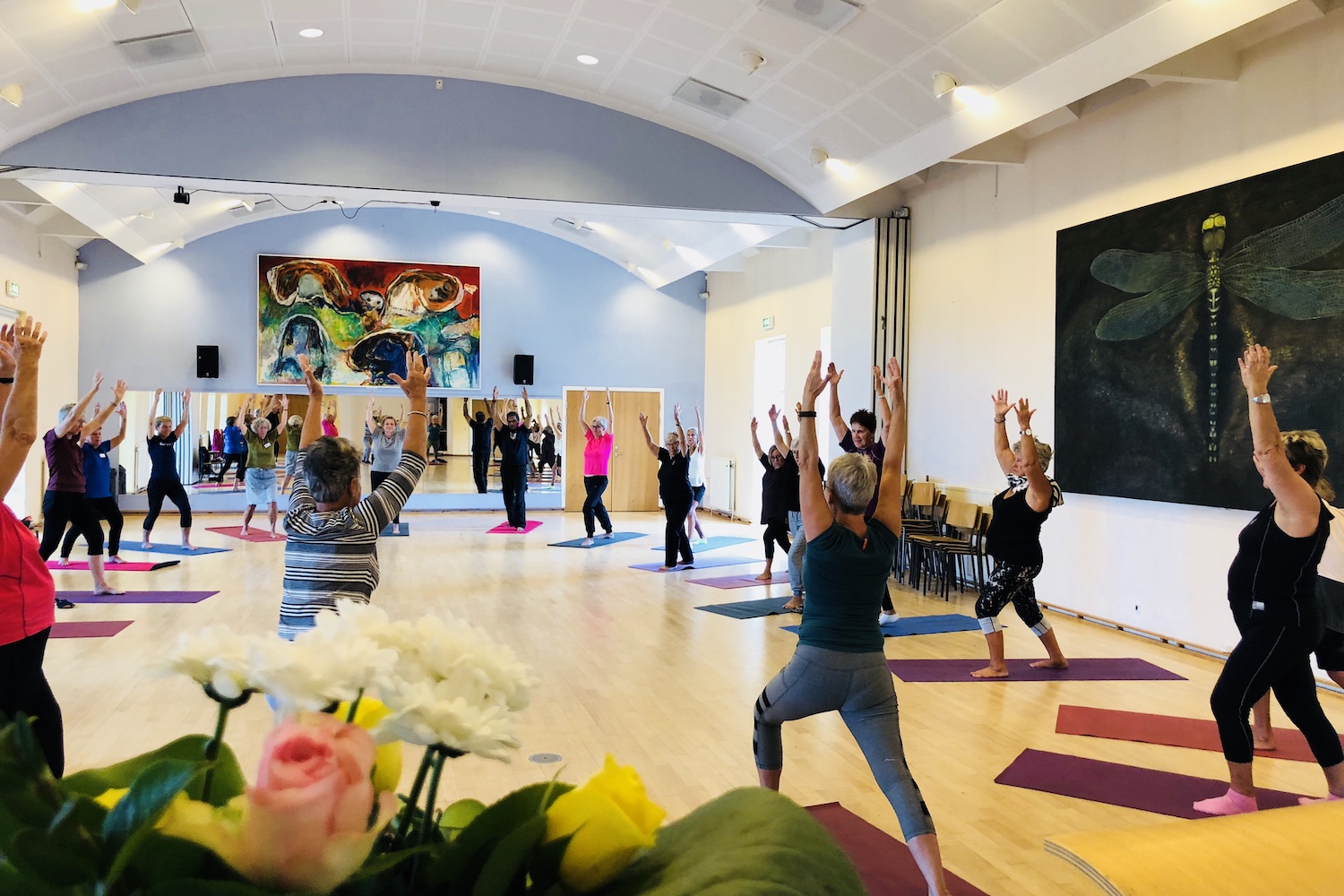 En gymnastiksal med en stor gruppe mennesker som står på yogamåtter med hænderne op i luften
